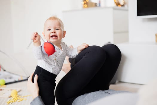 Happy family moments. Mother lying comfortably on children's mat watching and suppervising her baby boy playinghis in living room. Positive human emotions, feelings, joy.