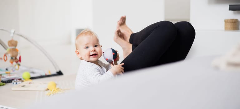 Happy family moments. Mother lying comfortably on children's mat playing with her baby boy watching and suppervising his first steps. Positive human emotions, feelings, joy