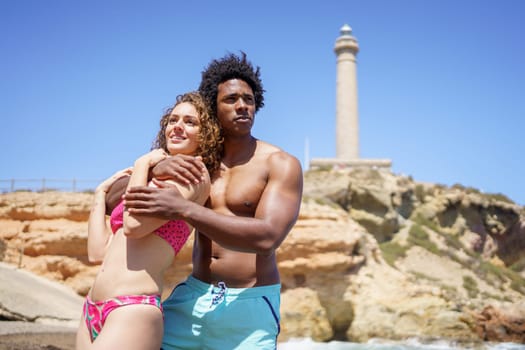 From below of African American boyfriend and girlfriend in pink bikini embracing each other while standing on seashore near lighthouse and looking away