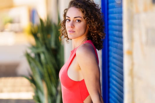 Side view of confident young female in red sundress with curly hair looking at camera, while standing against blue shutters and blurred tropical plants in daylight