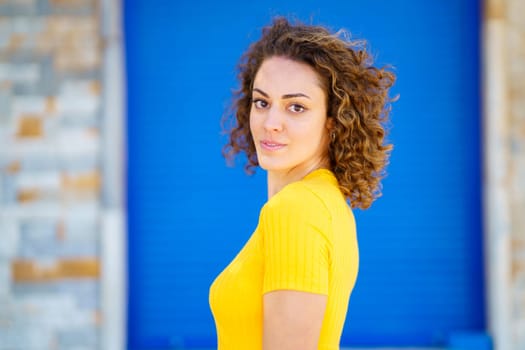 Side view of calm young female in yellow t shirt with curly brown hair looking at camera and smiling against blurred background on street