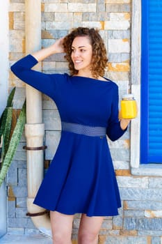 Positive young female in blue dress touching curly hair and looking away while standing on street with glass jar of orange juice against brick building