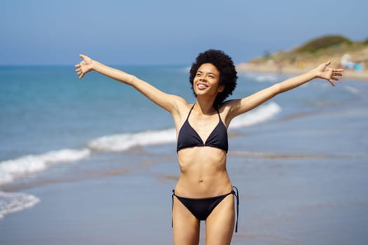 Positive African American female in swimwear with Afro hairstyle looking into distance with outstretched arms while standing on wet coast near sea