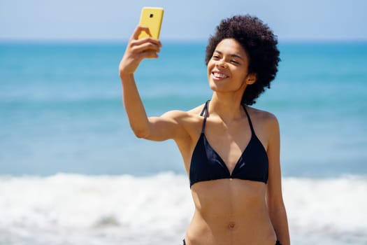Joyful African American female traveler in bikini taking self portrait on cellphone with smile while standing near waving sea in tropical resort