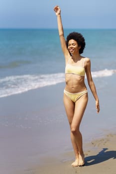 Full body of cheerful young African American female, traveler with dark curly hair in trendy bikini, smiling and looking away while walking on sandy beach with raised arm in sunlight