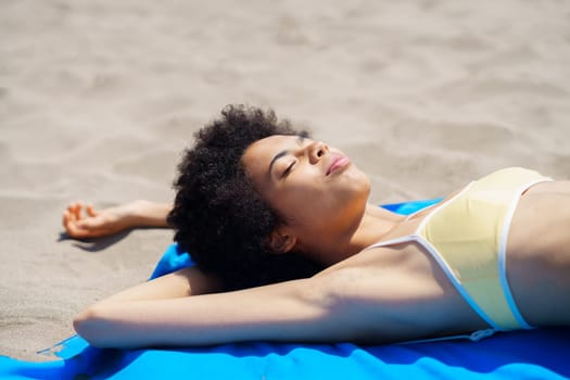 Happy young African American female tourist with curly hair in trendy bikini lying on sandy beach with closed eyes while sunbathing during summer holidays
