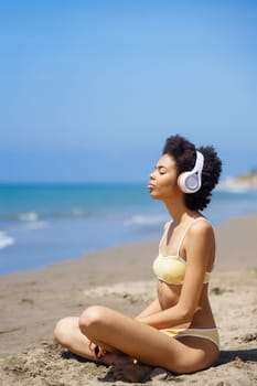 Side view of young fit ethnic female tourist with Afro hair in swimwear listening to music in headphones, while meditating on sandy beach with closed eyes in Lotus pose