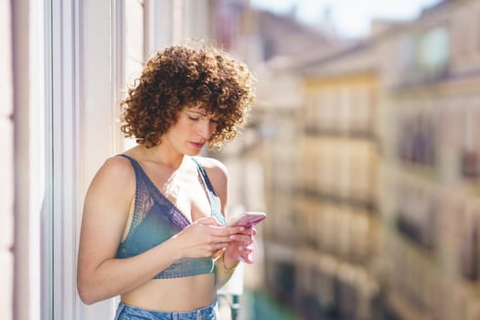 Positive young female in bra and jeans with curly hair while browsing smartphone on sunny balcony