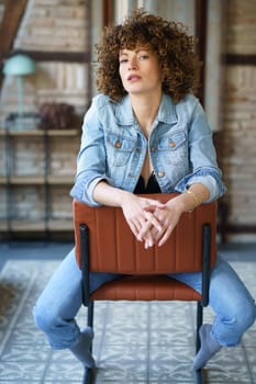 Serious young woman wearing denim jacket sitting on leather chair and touching hair while looking at camera against blurred brick wall