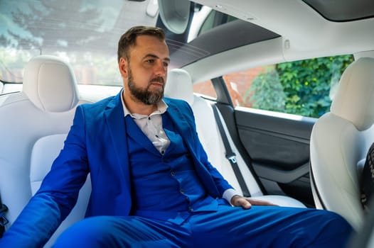 A caucasian man in a blue suit sits in the back seat of a car. Business class passenger