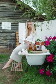 A woman is sitting on a cast-iron bathtub in the courtyard of a country house next to a bush of flowering peonies. The concept of summer, country life, a bathroom on the street in a blooming garden in the country