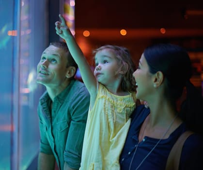 Family, aquarium and child pointing at fish for learning, curiosity or knowledge, bonding or care. Mother, fishtank and happy kid with father watching marine animals swim underwater in oceanarium