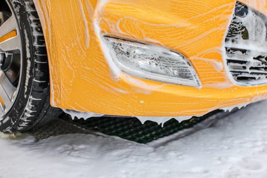 Yellow car front and light washed in self serve carwash. Bumper covered with white shampoo foam.