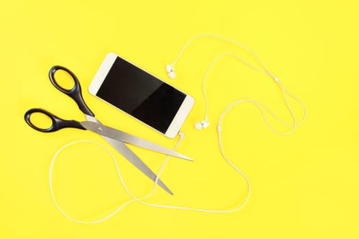 Flat lay photo, white mobile phone with ear buds , scissors about to cut the cable. Yellow background.