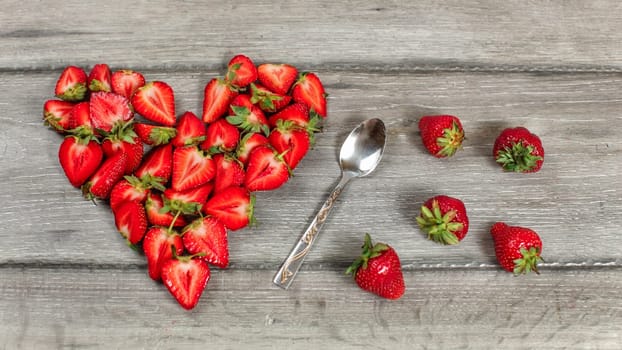 Top down view, strawberry pieces arranged in shape of heart, spoon and some spilled strawberries near, on gray wood desk.