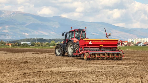 Red tractor pulling sowing trailer behind over empty field. Some village and mountains in background.