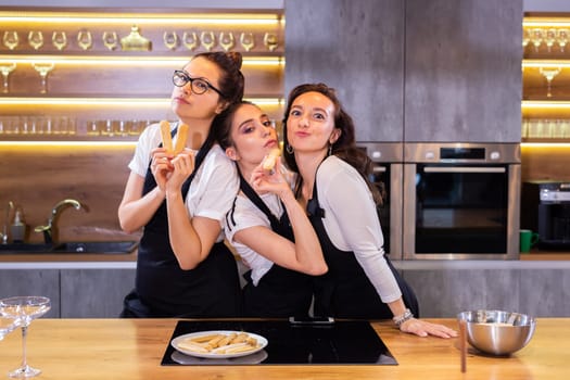 Three funny female chef in uniform holding cookies while smiling and having fun at camera in kitchen