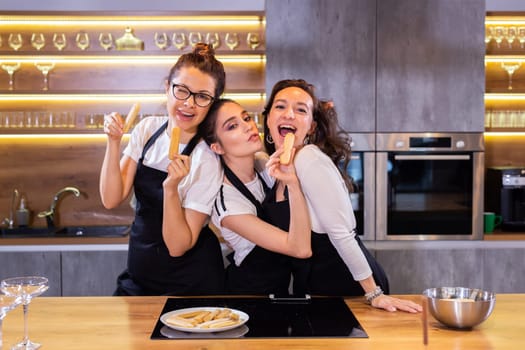 Three funny female chef in uniform holding cookies while smiling and having fun at camera in kitchen