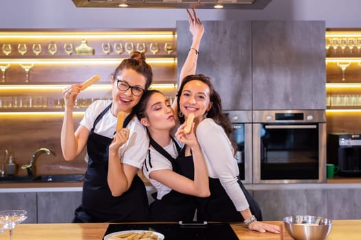 Three funny female chef in uniform holding cookies while smiling and having fun at camera in kitchen