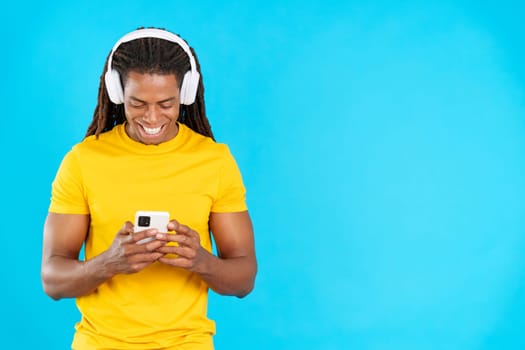 Latin man with dreadlocks listening to music with headphones and a mobile in studio with blue background