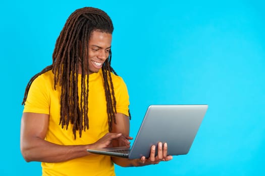 Latin man with dreadlocks smiling while using a laptop in studio with blue background