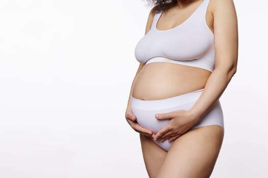 Close-up studio shot of a pregnant woman, holding her belly, posing in white lingerie over isolated background with copy ad space. Nudity. Body positivity. Natural beauty people. Pregnancy. Maternity