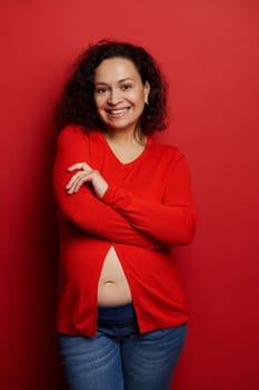 Happy multi ethnic pregnant woman smiling looking at camera, posing with folded arms over red color background. Vertical studio portrait of gravid female, expectant mother. Carefree pregnancy 24 week