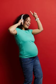 Happy positive pregnant woman in casual clothes, wearing wireless headphones, having fun, dancing, singing, listening to music, isolated on red background. Pregnancy. Maternity. Lifestyle