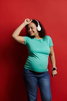 Adult pregnant woman wearing casual clothes, dancing while listening to music on wireless headphones, enjoying wonderful moments of happy carefree pregnancy, isolated on red background. Maternity