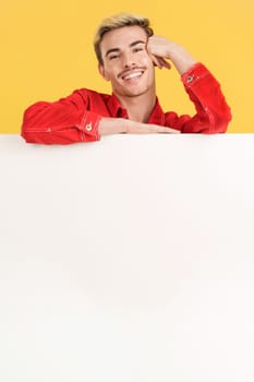 Cheerful gay man leaning on a blank board in studio with yellow background