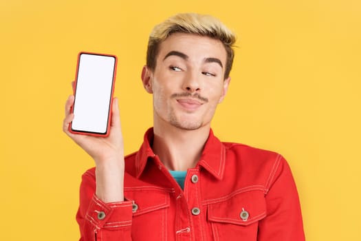 Happy gay man holding a mobile with a blank screen in studio with yellow background