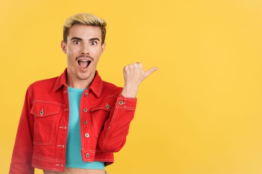 Surprised gay man pointing a blank space to the side in studio with yellow background