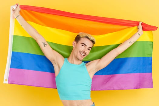 Proud gay man raising a lgbt rainbow flag in studio with yellow background