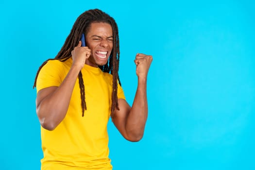 Latin man with dreadlocks celebrating while talking to the mobile in studio with blue background