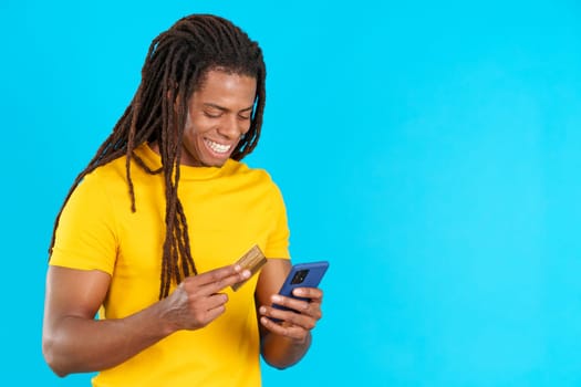 Happy latin man with dreadlocks using mobile and card to shopping online in studio with blue background
