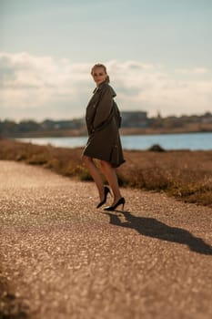 Portrait blonde sea cape. A calm young blonde in an unbuttoned khaki raincoat walks along the seashore, under a raincoat a black skirt and top.