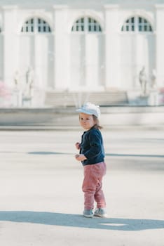 A female toddler in a cap and velvet overall with her back turns in the yard of the European palace. A baby girl walks to the fountain.