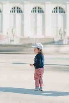 A female toddler in a cap and velvet overall with her left side in the yard of the European palace. A baby girl walks to the fountain.