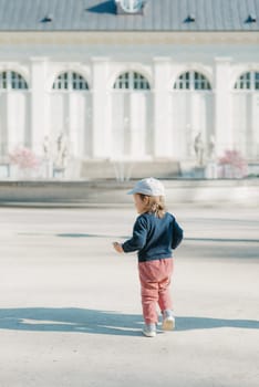 A female toddler in a cap and velvet overall with her back in the yard of the European palace. A baby girl walks to the fountain.