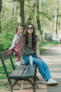 A mother rests close to her little daughter on the bench in the park. A female toddler in velvet overall is having fun with her mom in the garden.