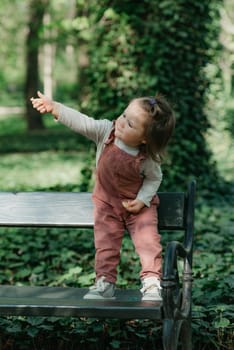 A female toddler in a velvet overall holds out a cookie on the garden bench. A baby girl is having fun in the park.