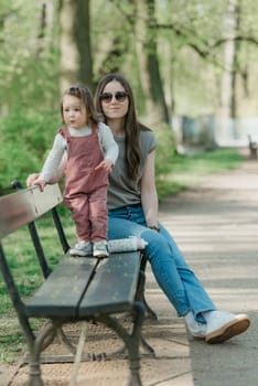 A mother rests close to her little daughter on the bench in the green park. A female toddler in velvet overall is having fun with her mom in the garden.