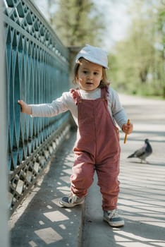 A female toddler eats a sausage close to the garden bridge fence. A baby girl in a cap and velvet overall walks near the pigeon in the park.