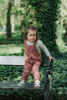 A female toddler in a velvet overall holds is on the garden bench. A baby girl with a small ponytail is having fun in the park.