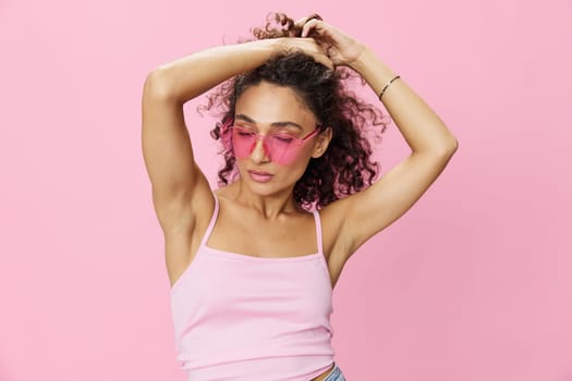 Happy woman hands up waxing her armpits, shaving, with curly hair in a pink tank top and jeans on a pink background wearing sunglasses with a nice tan, copy space. High quality photo