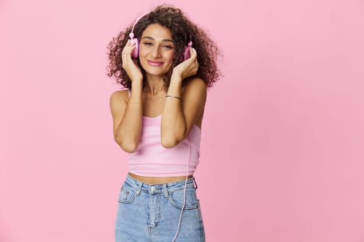 Happy woman wearing headphones with curly hair listening to music and singing along with her eyes closed in a pink T-shirt and jeans on a pink background, copy space. High quality photo