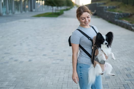 Happy caucasian woman walking with a dog in a backpack. Papillon Spaniel Continental in a sling