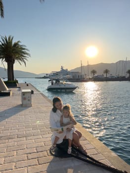 Mom reads a book to a little girl sitting on a bollard on a pier by the sea. High quality photo