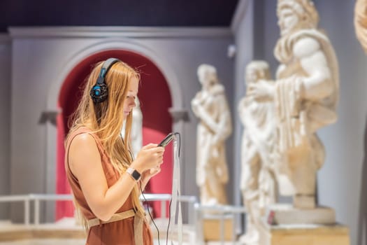 Portrait of contemporary young woman looking at sculptures and listening to audio guide at museum exhibition.