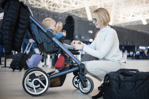 Motherat interacting with her infant baby boy child in stroller while travelling at airport terminal station. Travel with child concept